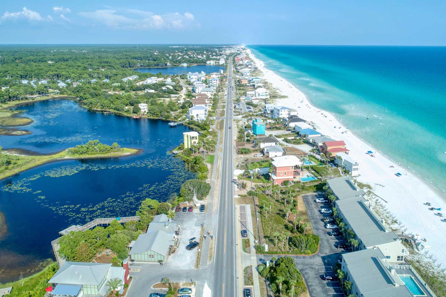 Aerial view of Florida’s Highway 30A running straight between a deep blue coastal dune lake on the left and a long stretch of bright white-sand beach and clear turquoise Gulf water on the right, with colorful beach houses and low-rise condos lining both sides of the road under a clear blue sky.