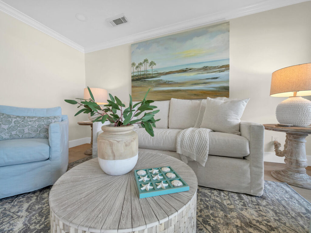 Soft neutral living room with a light beige sofa, pale blue armchair, round wooden coffee table, coastal decor, and a large landscape painting of dunes and palm trees hanging on the wall behind.