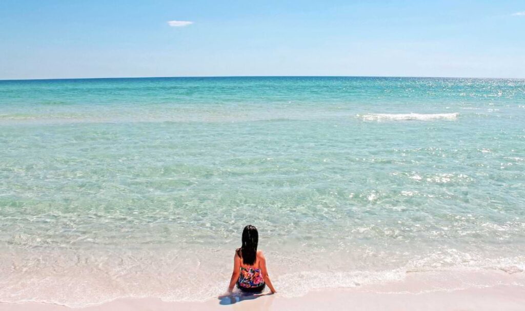 Child sitting at the edge of clear, shallow turquoise water on a white-sand beach, facing the open sea under a bright blue sky.