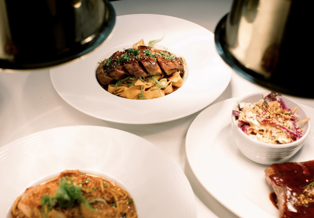 Overhead view of a white table set with a main dish of sliced glazed meat over wide noodles, accompanied by colorful slaw and other entrées, illuminated by warm pendant lights at Aja.