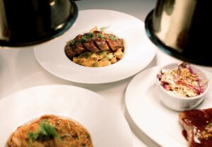 Overhead view of a white table set with a main dish of sliced glazed meat over wide noodles, accompanied by colorful slaw and other entrées, illuminated by warm pendant lights at Aja.