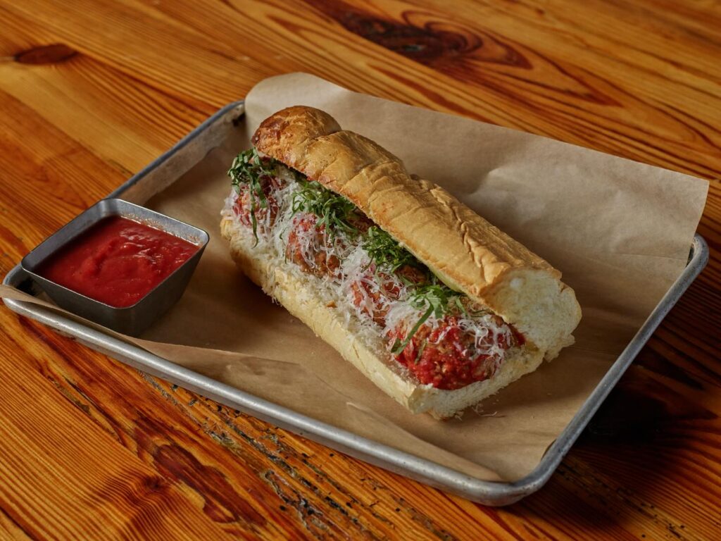 Close-up of a toasted meatball sub loaded with marinara sauce, shredded cheese, and fresh herbs on a hoagie roll, served on parchment paper with a side of red dipping sauce at Amici.