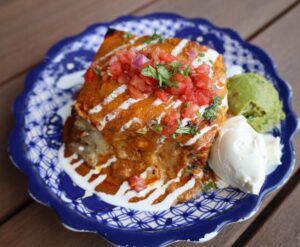 Hearty layered enchilada-style casserole topped with diced tomatoes and cilantro and drizzled with white sauce, served on a blue patterned plate with scoops of guacamole and sour cream at Amigos 30A.