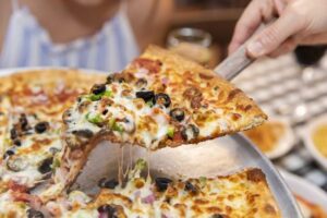 Close-up of a hand lifting a cheesy slice of supreme pizza loaded with olives, peppers, and other toppings from a metal pan, with melted cheese stretching from the remaining slices at Angelina’s.