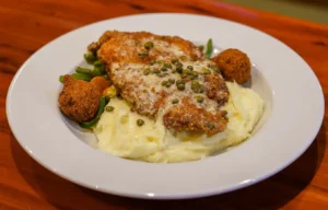 Breaded chicken cutlet topped with creamy caper sauce, served over a bed of mashed potatoes with green beans and two hushpuppies on a white plate.