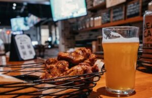 Basket of crispy chicken wings and a pint of hazy golden beer on a wooden bar, with TVs and taproom decor softly blurred in the background at Beach Camp Brewing Company.