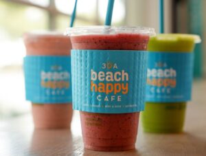 Three bright smoothies in clear to-go cups wrapped with blue “30A Beach Happy Café” sleeves, with pink and green drinks lined up on a counter and blue straws poking through the lids.