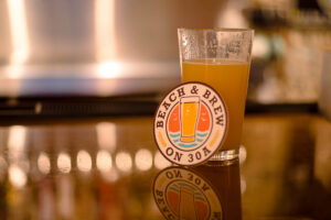 Close-up of a hazy golden beer in a pint glass on a shiny bar top, with a round “Beach & Brew on 30A” logo coaster propped in front, reflecting warmly in the counter’s surface.