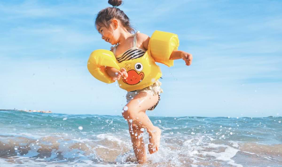 Young child in a striped swimsuit and bright yellow duck-themed floaties splashing joyfully in shallow ocean waves under a clear blue sky.