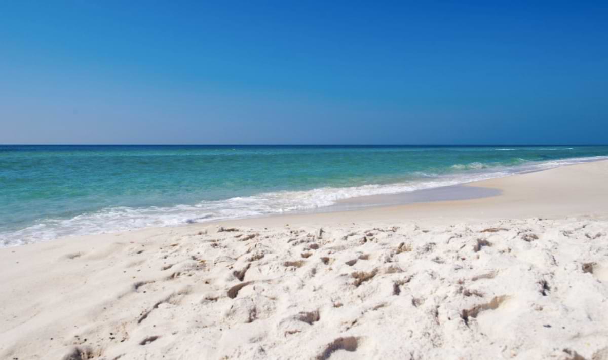 Pristine white-sand beach with soft footprints leading to gentle turquoise waves, under a clear blue sky stretching across the horizon.