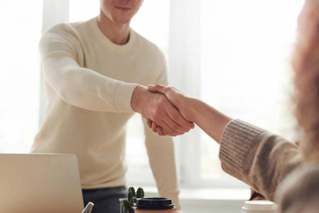 Close-up of two people reaching across a desk to shake hands in a bright office, symbolizing choosing a vacation rental manager, with a laptop and coffee cups visible in the foreground.