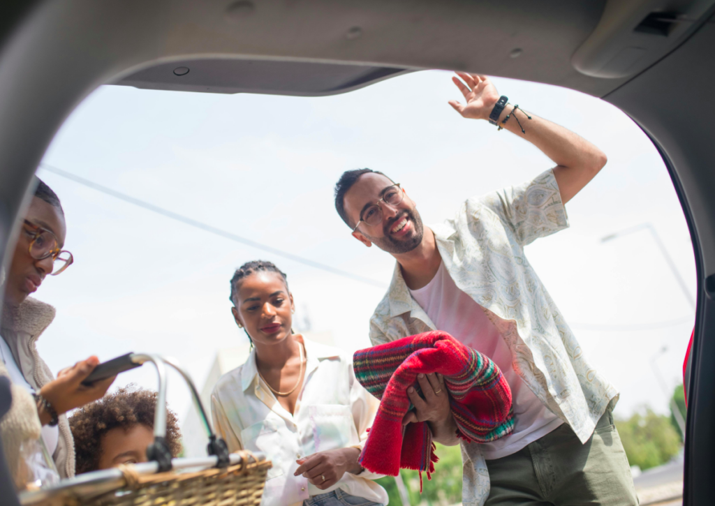 View from inside an open car trunk looking out at a group of friends loading items for a trip, including a picnic basket and colorful blanket, as one person waves and leans toward the car.