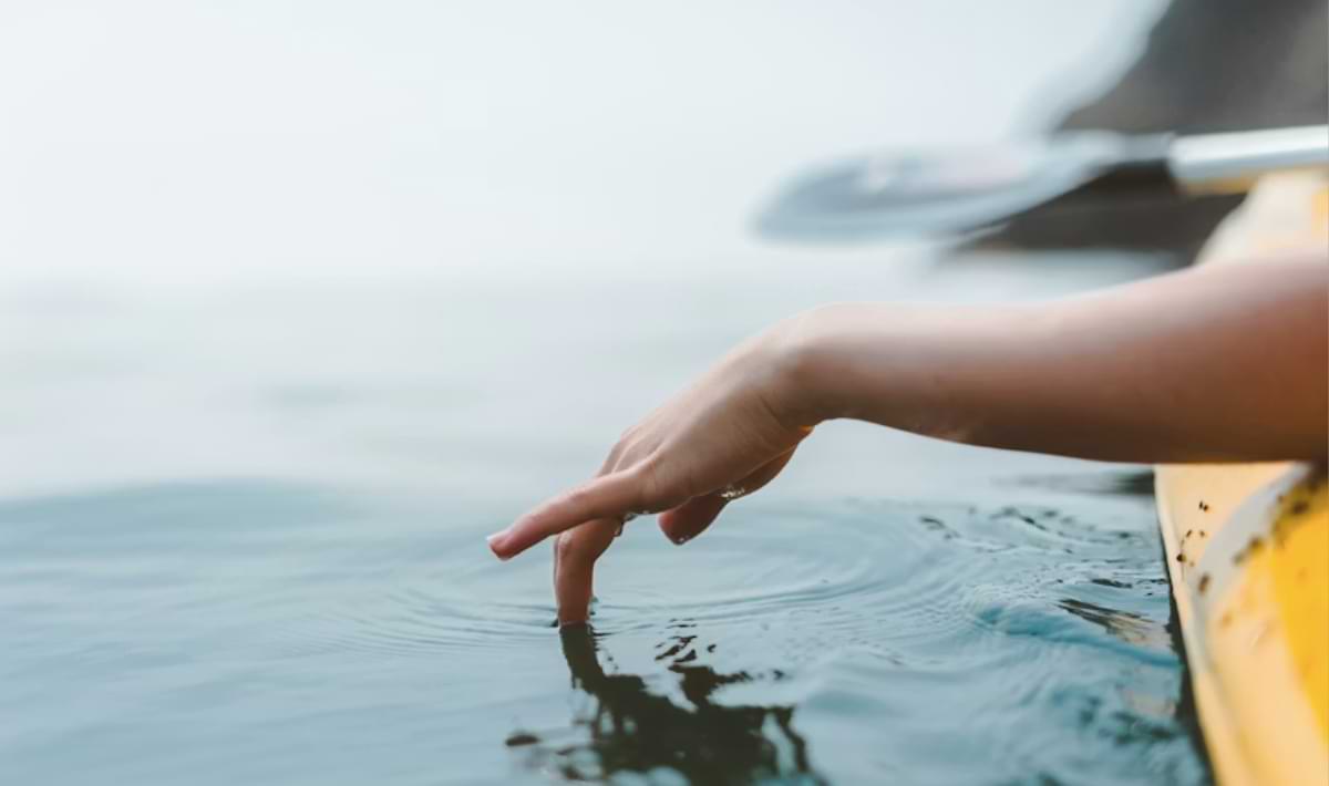 Close-up of a person’s hand trailing in calm water, fingertips creating soft ripples beside a yellow kayak on a hazy, peaceful day.