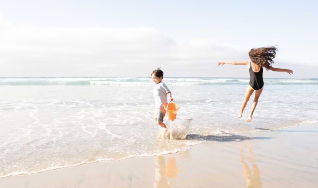 Two children playing at the shoreline, one running with an orange bucket as waves splash around their feet and the other in a black swimsuit leaping joyfully above the wet sand, with gentle surf in the background.