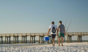 Two people walk side by side along a white-sand beach carrying fishing rods and a blue bucket, heading toward the shoreline beside a wooden pier extending over the Gulf of Mexico under a clear blue sky.