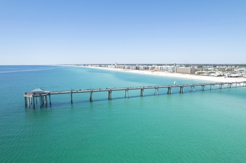 A long concrete fishing pier stretches far out over calm, bright turquoise Gulf water toward the left, with people walking along it and a small covered pavilion at the pierhead, while a distant line of white-sand beach and low-rise condos curves along the shoreline under a cloudless blue sky.