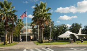 Front of a palm-lined airport entrance with an American flag flying and a displayed fighter jet on the lawn under a bright blue sky with scattered clouds.