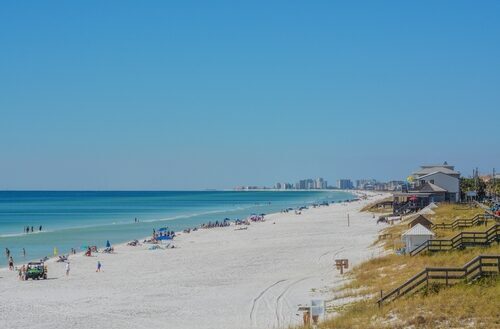 View down a long stretch of Miramar Beach with calm turquoise water on the left, scattered beachgoers and umbrellas on wide white sand, and a line of houses and walkovers along the dunes with a cluster of high-rise condos visible in the distant background under a clear blue sky.