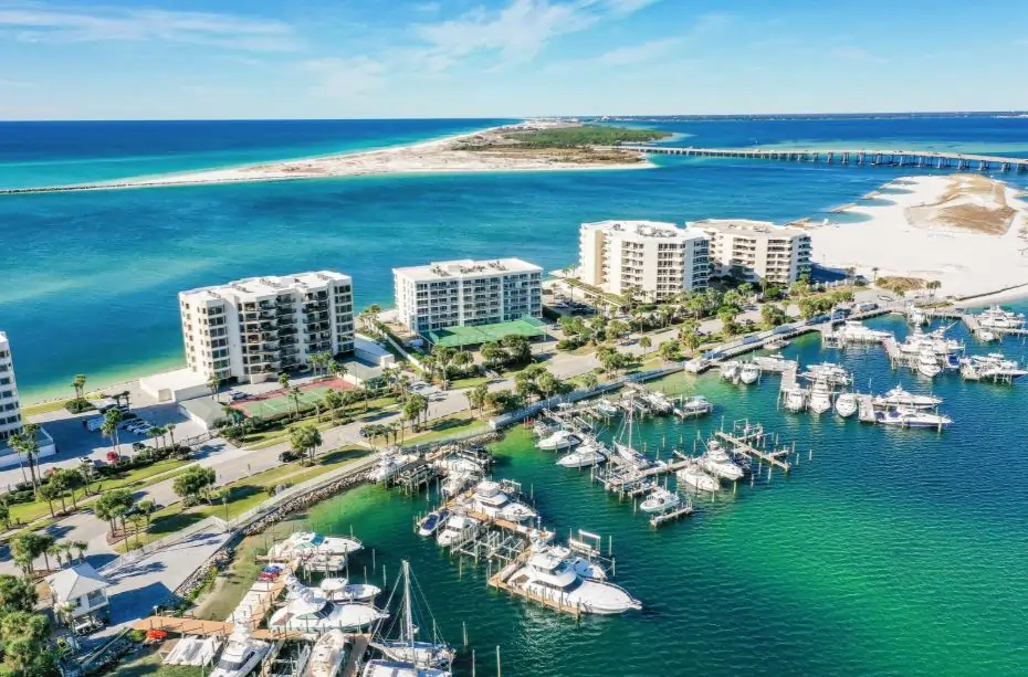 Aerial view of a coastal resort area with waterfront condo buildings, a busy marina filled with boats, and a narrow sandbar and bridge stretching across clear blue-green water.