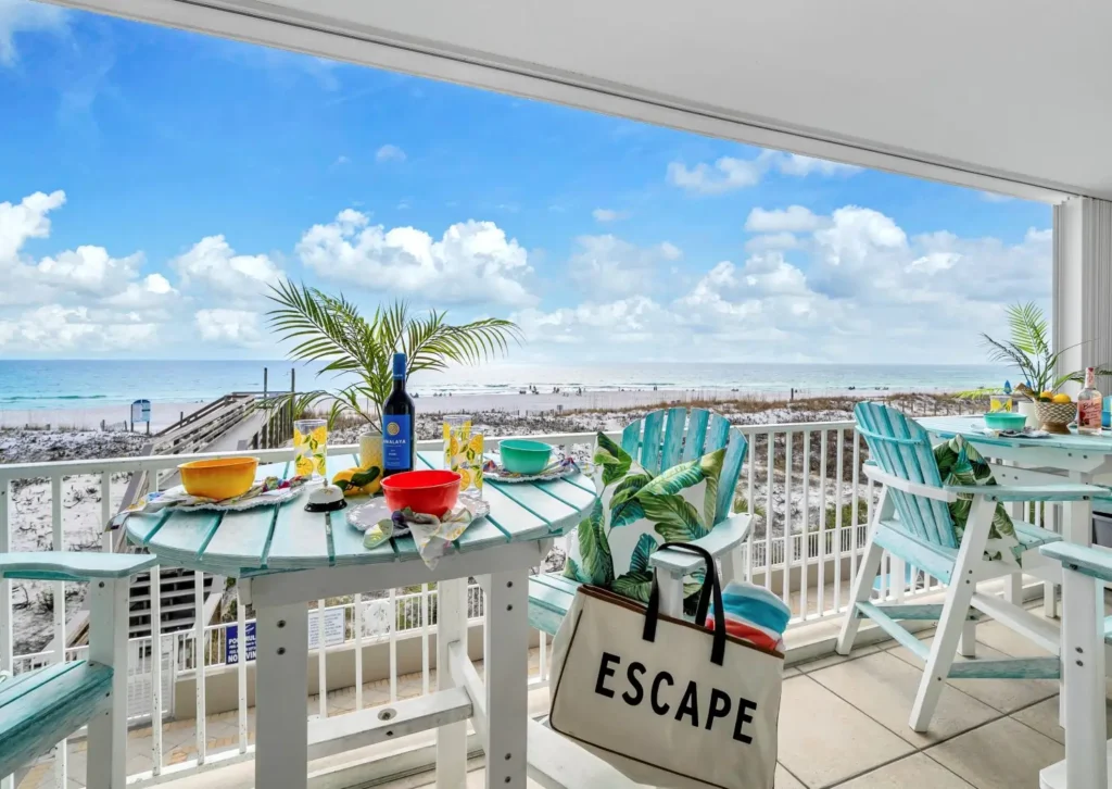 Covered balcony with pastel blue Adirondack chairs and tropical cushions around café tables set for a meal, overlooking a boardwalk, white-sand beach, and turquoise gulf under a bright blue sky with puffy clouds.