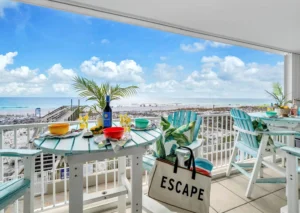 Covered balcony with pastel blue Adirondack chairs and tropical cushions around café tables set for a meal, overlooking a boardwalk, white-sand beach, and turquoise gulf under a bright blue sky with puffy clouds.