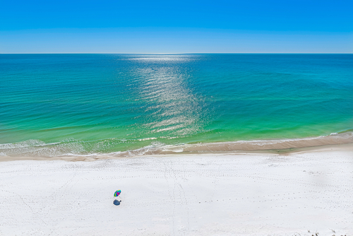 Minimalist view of Panama City Beach with gentle emerald-green waves rolling onto a wide, empty stretch of sugar-white sand, two colorful beach umbrellas set up near the surf, and bright sunlight shimmering across the calm Gulf under a clear blue sky.