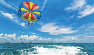 Two people sit side by side in harnesses under a bright multicolored parasail, soaring high above choppy deep-blue Gulf water behind a speedboat whose white wake cuts across the foreground, with a distant strip of coastline and puffy white clouds under a vivid blue sky.