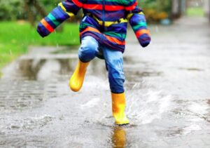 Child in a colorful striped raincoat and bright yellow boots joyfully jumping into a puddle on a wet sidewalk, splashing water in the rain.