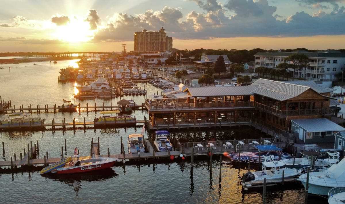 Sunset view of Destin Harbor with a busy waterfront restaurant on stilts, rows of docked boats, and the sun glowing over the marina and high-rise condos along the Emerald Coast.