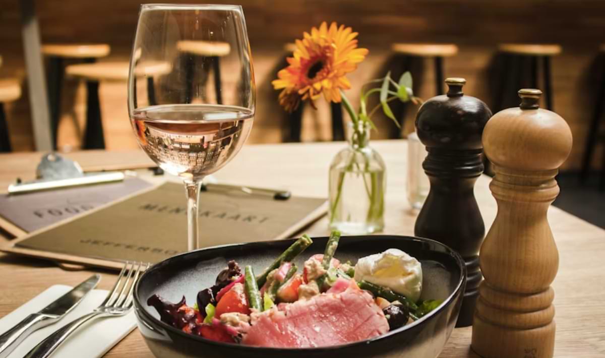 Close-up of a restaurant table set with a glass of rosé wine, a black bowl of colorful gourmet salad, salt and pepper mills, and a single orange flower in a vase, with menus blurred in the background.