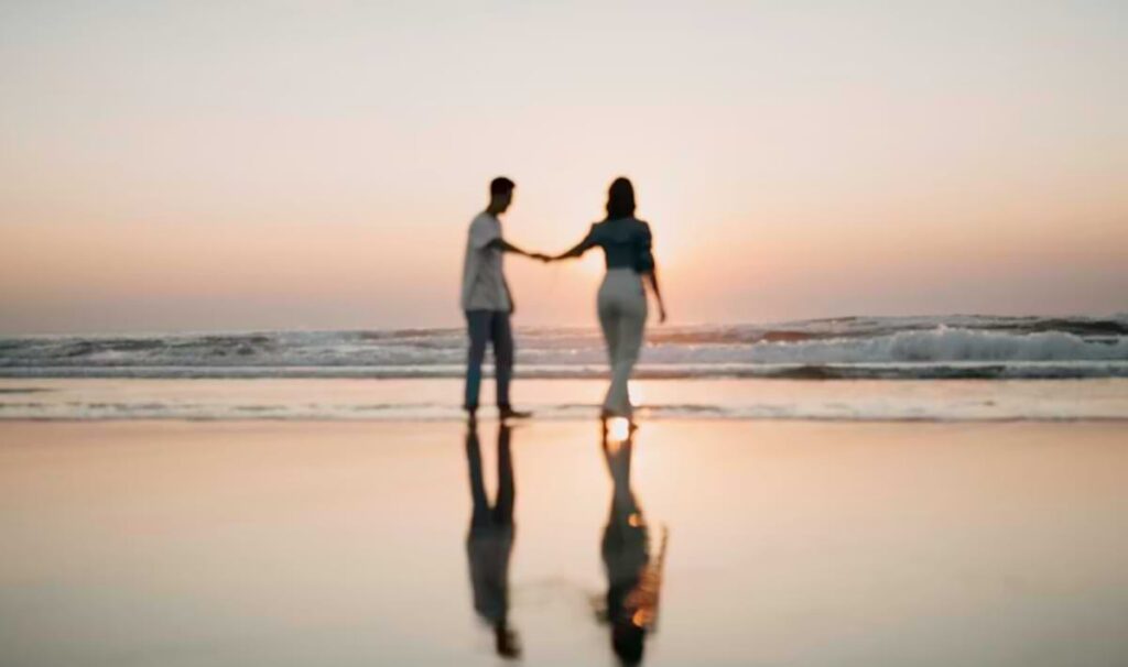 Silhouetted couple holding hands and walking along the shoreline at sunset, reflected in the wet sand with gentle waves rolling in behind them.