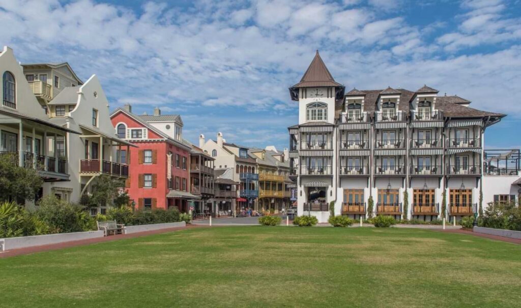 Colorful European-style buildings and a tall, turreted hotel facing a central green lawn in Rosemary Beach, with balconies, shops, and cafes lining the pedestrian-friendly square under a partly cloudy sky.