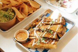Overhead view of a metal tray filled with folded quesabirria-style tacos cut into wedges, topped with crema and chopped cilantro, served with a small cup of orange dipping consomé, alongside another tray holding tortilla chips and a paper cup of guacamole.