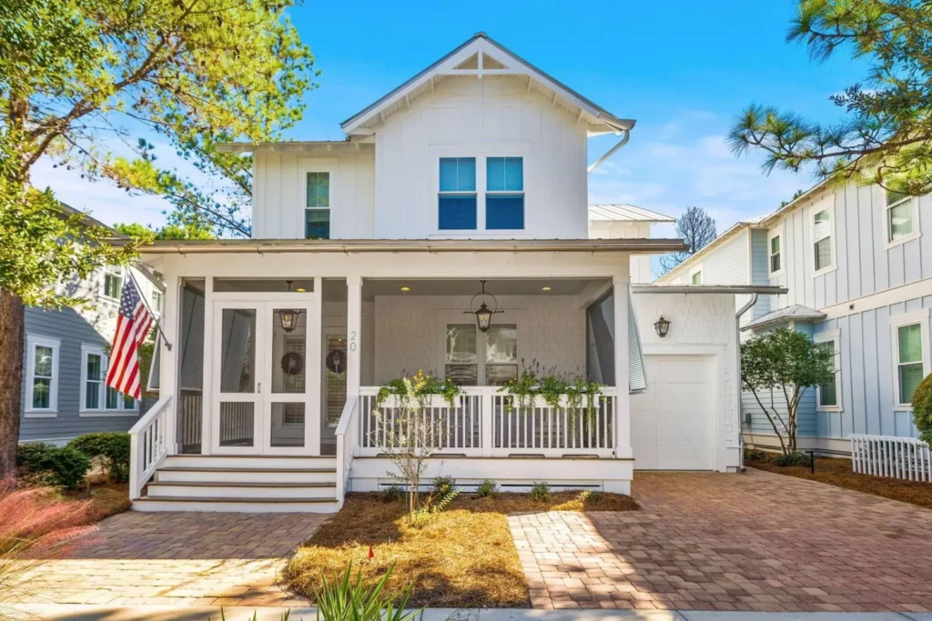 Bright white two-story coastal cottage with a screened front porch, American flag, and brick driveway, framed by trees and neighboring pastel homes under a clear blue sky.