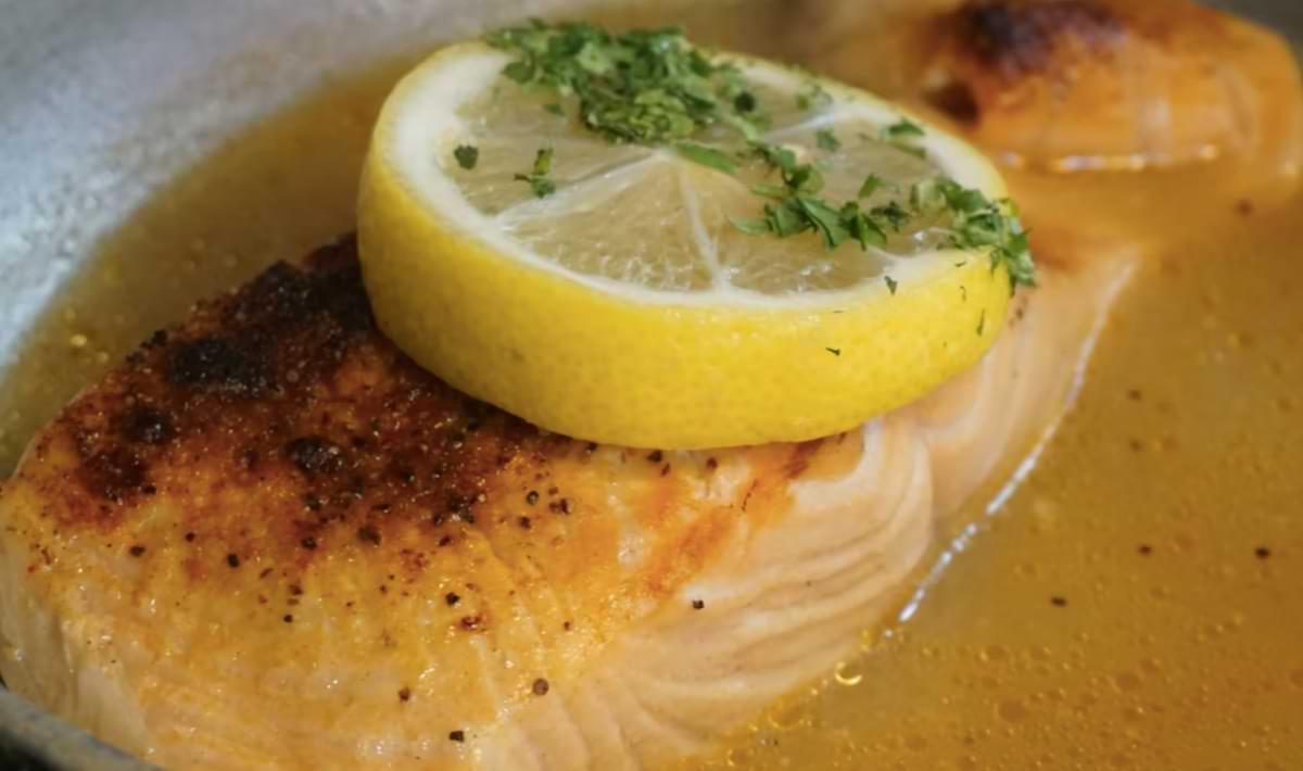 Close-up of a seared salmon fillet simmering in a golden butter sauce, topped with a thick lemon slice and sprinkled with fresh herbs at a Destin and Okaloosa Island seafood spot.