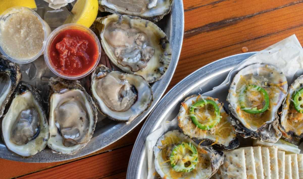Overhead view of two metal trays filled with raw oysters on the half shell with cocktail and horseradish sauces, and baked oysters topped with melted cheese and jalapeño slices, served alongside saltine crackers and lemon wedges on a wooden table.