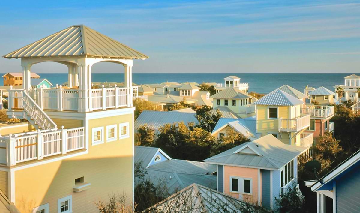 Colorful beach cottages with metal roofs and wraparound porches clustered along the coast, with a tall yellow home featuring a rooftop lookout tower in the foreground and the Gulf visible in the distance.