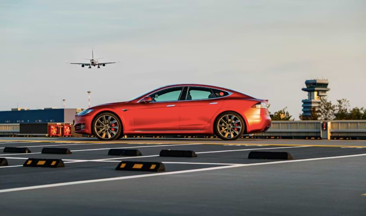 Side view of a shiny red sedan parked on an airport rooftop with a plane coming in for landing in the background and the control tower visible to the right.