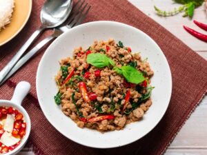 Overhead view of a white bowl filled with Thai basil stir-fried minced meat and red chilies, garnished with fresh basil leaves, set on a woven placemat with a side dish of chili fish sauce and silverware nearby.