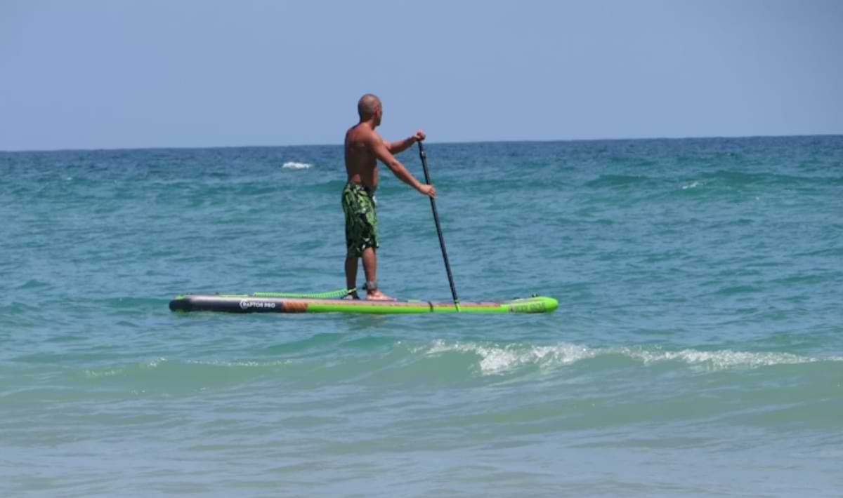 Man stand-up paddleboarding on a green board over gentle turquoise waves, facing the open water under a clear blue sky.