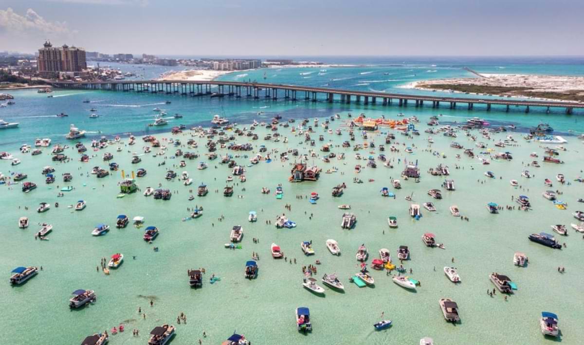 Aerial view of a crowded sandbar in clear turquoise water, packed with anchored boats and people wading and swimming, with a long bridge and city shoreline in the background.