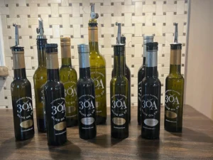 Assortment of dark and green glass bottles of 30A Olive Oil Co. products arranged on a wooden counter, each labeled with different infused olive oils and balsamic vinegars, set against a tiled kitchen backsplash.