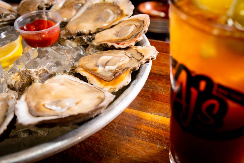 Tray of raw oysters on ice with cocktail sauce and lemon wedges next to a cold drink in an AJ’s logo glass on a wooden table.