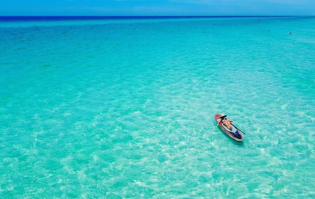 Person lying back and sunbathing on a paddleboard floating alone in crystal-clear, shallow turquoise water that stretches to the horizon under a bright blue sky along Florida’s 30A coast.