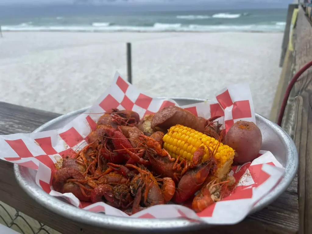 Tray of seasoned crawfish, corn on the cob, sausage, and a red potato on a beachside deck overlooking white sand and rolling waves.