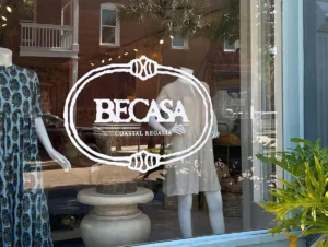 Shop window of Becasa boutique displaying two mannequins in casual coastal dresses, with the white “BECASA Coastal Regalia” logo printed on the glass and reflections of brick buildings and palm trees outside.