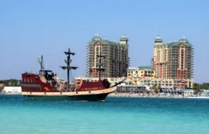 Large red and cream pirate-themed tour boat named “Buccaneer” cruising through clear turquoise water with tall resort towers and waterfront buildings of Destin Harbor rising in the background under a clear blue sky.