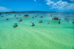 Aerial view of Crab Island with dozens of boats anchored in shallow, clear turquoise water, people swimming and wading between them, and a floating tiki-style structure under a bright blue sky with scattered clouds.