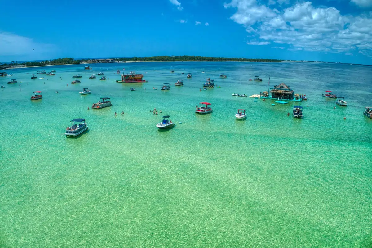 Aerial view of Crab Island with dozens of boats anchored in shallow, clear turquoise water, people swimming and wading between them, and a floating tiki-style structure under a bright blue sky with scattered clouds.