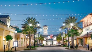 View down the main street of Destin Commons at dusk, with palm trees wrapped in lights, strings of glowing bulbs overhead, and storefronts like Sephora lining the outdoor shopping promenade under a clear blue sky.